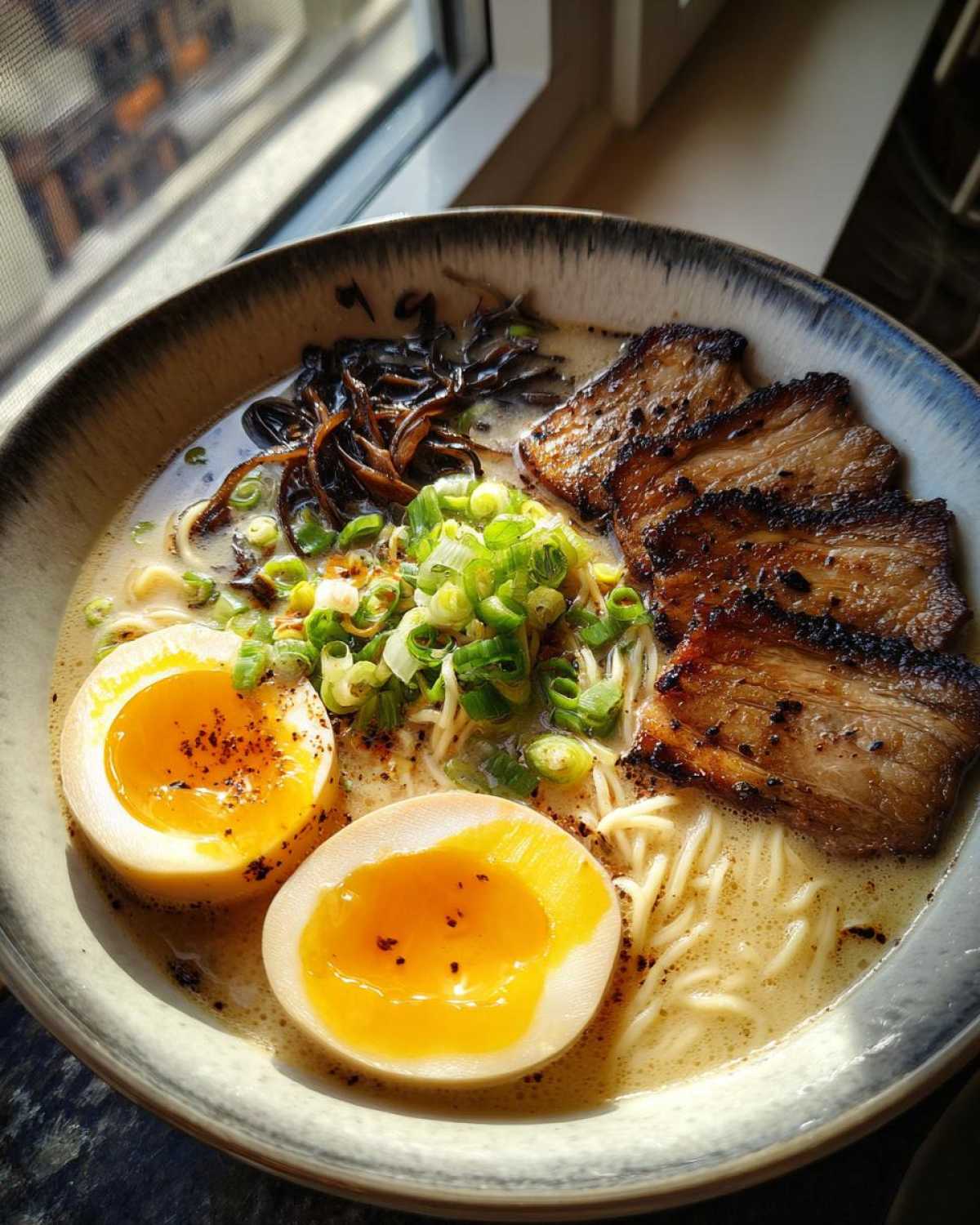 A close-up bowl of Tonkotsu Ramen featuring creamy broth, noodles, seared chashu pork, ajitama eggs, and scallions.