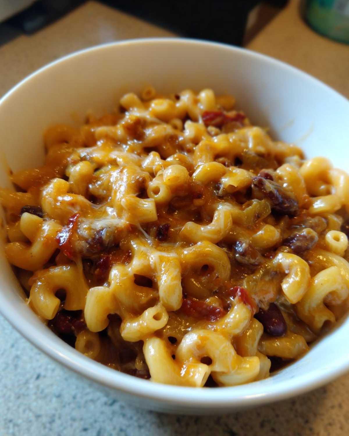 A close-up shot of a white bowl filled with creamy Vegetarian Chili Mac, topped with melted cheddar cheese and visible kidney beans.
